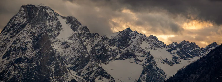 Snowy mountain peaks under dark clouds with warm light breaking through near the horizon.