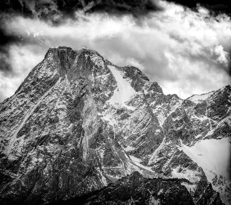 Black-and-white close view of a steep, snow-streaked mountain beneath textured clouds.
