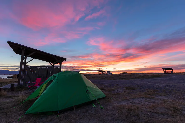 A green tent stands beside a wooden shelter as pink clouds spread across the sunset horizon.
