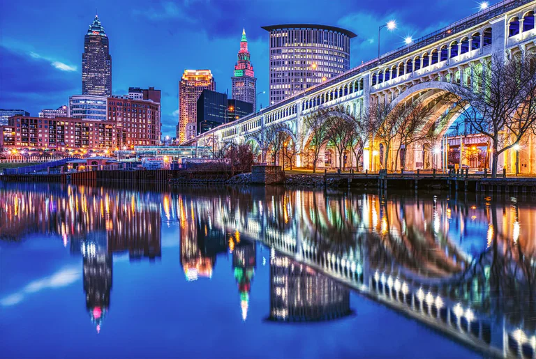 Lit bridge arches and downtown Cleveland buildings reflected in a river at night.