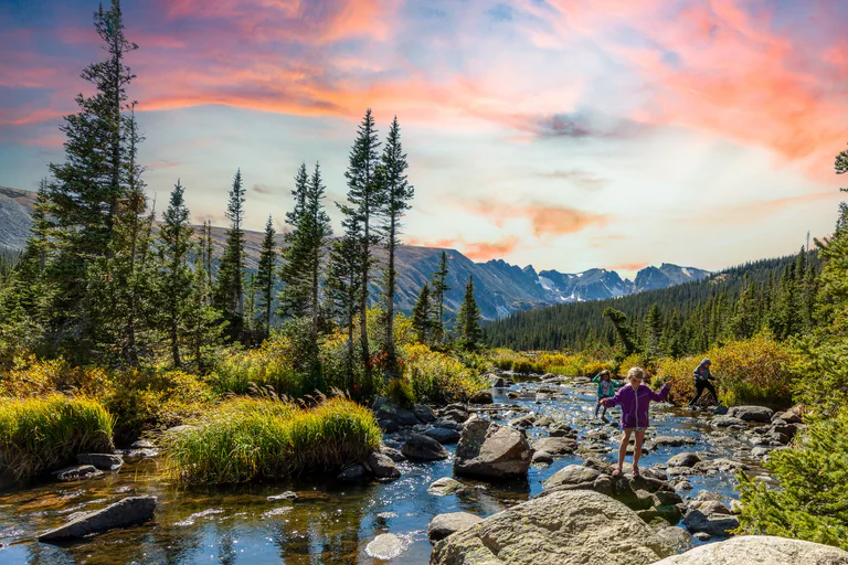 A person stands among rocks in a shallow mountain stream beneath conifers and a pink sky.