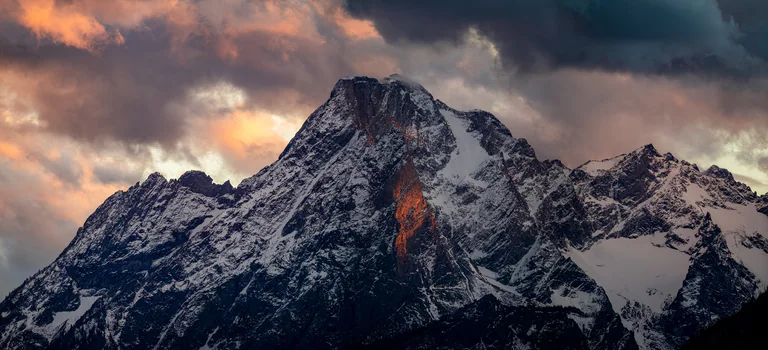 Warm light catches part of a rugged snowy mountain range beneath gray clouds.