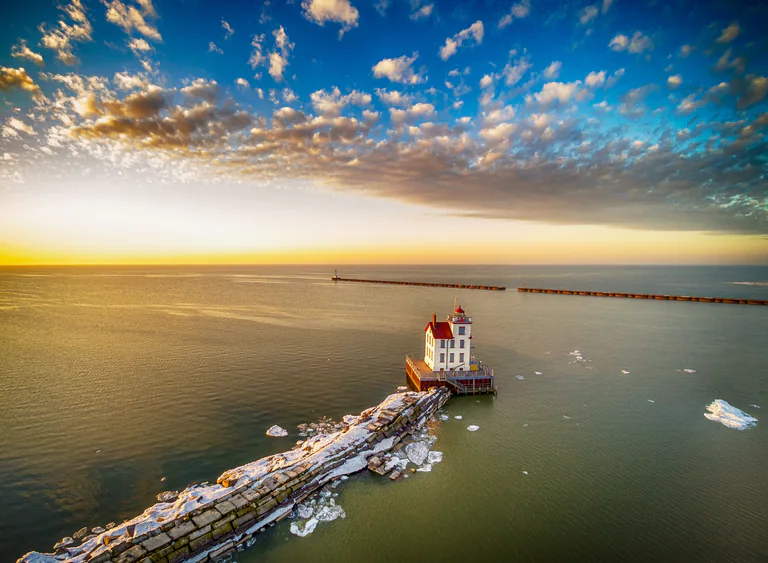 Aerial view of a long breakwater leading to a lighthouse in calm water at sunset.