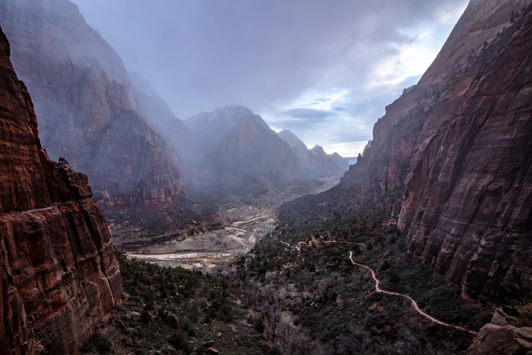 Low clouds and mist soften red cliffs above a green canyon floor and winding river.
