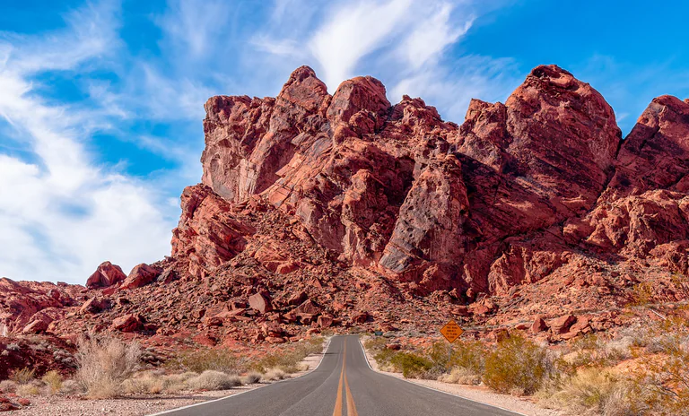 A paved road winds beneath jagged red sandstone formations under wispy white clouds.