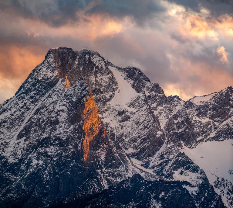 A shaft of warm light illuminates a steep snowy mountain face beneath a cloudy sky.