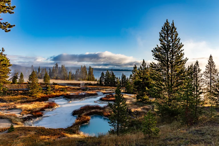 A stream runs between rocky banks and conifers, with distant mountains under a blue sky.