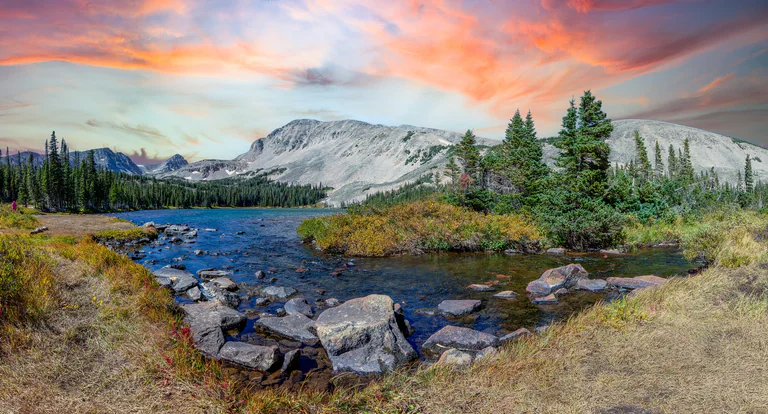A rocky shoreline and conifers beside blue water, with pale mountains under a pink sky.