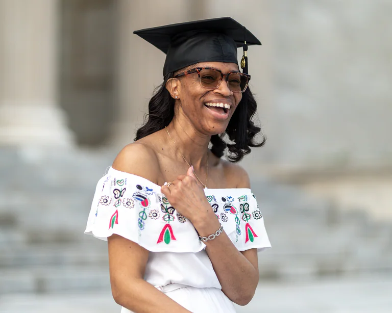 A woman in a black graduation cap and embroidered white top laughs while looking to one side.