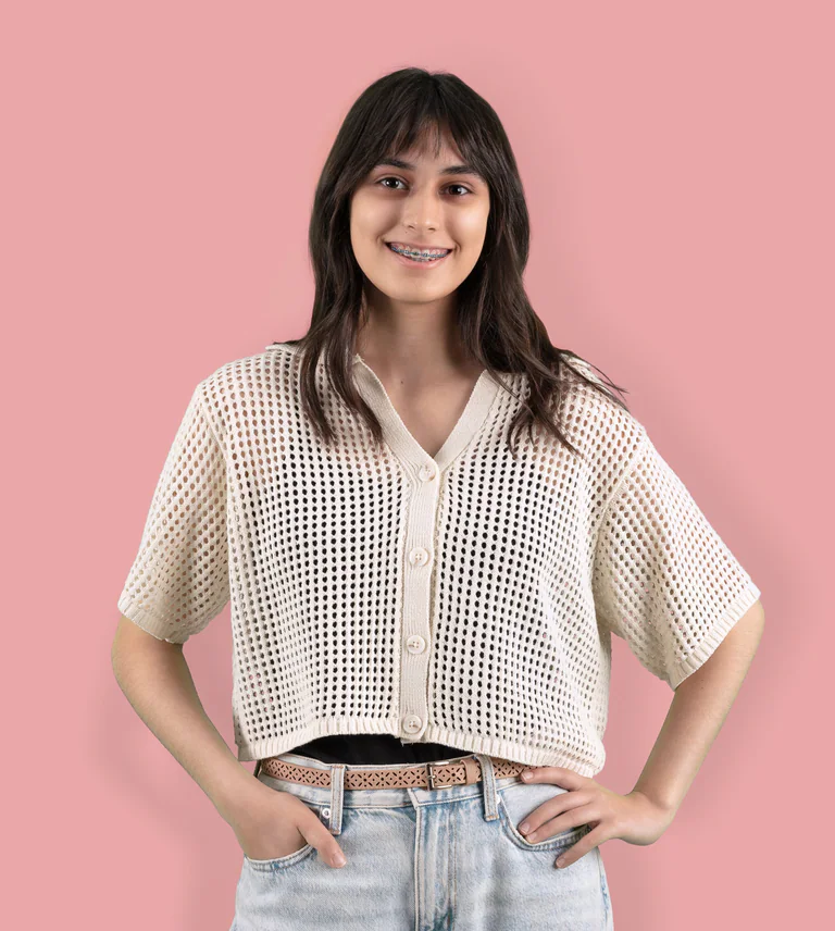 A smiling woman in a white knitted top poses with her hands on her hips against a pink background.