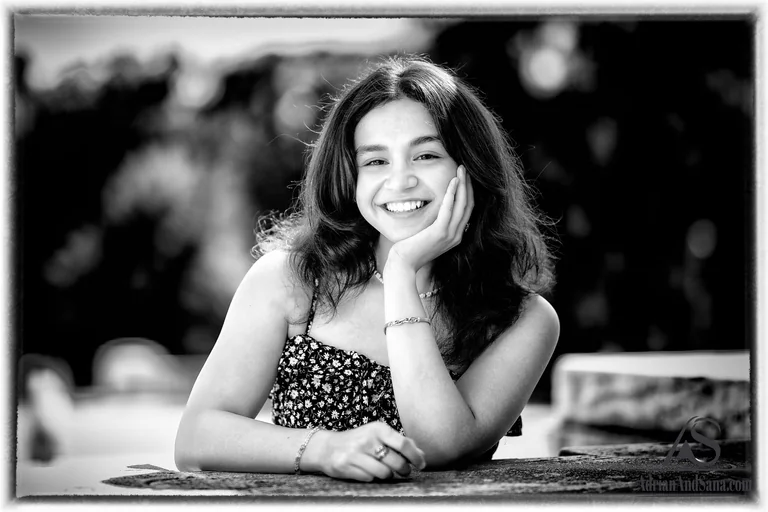 Black-and-white portrait of a smiling woman resting her cheek on one hand at an outdoor table.