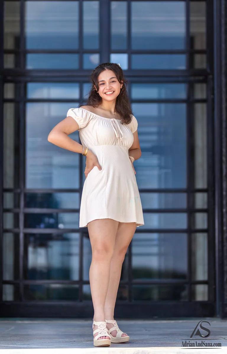Full-length portrait of a woman in a white dress standing in front of tall glass doors.