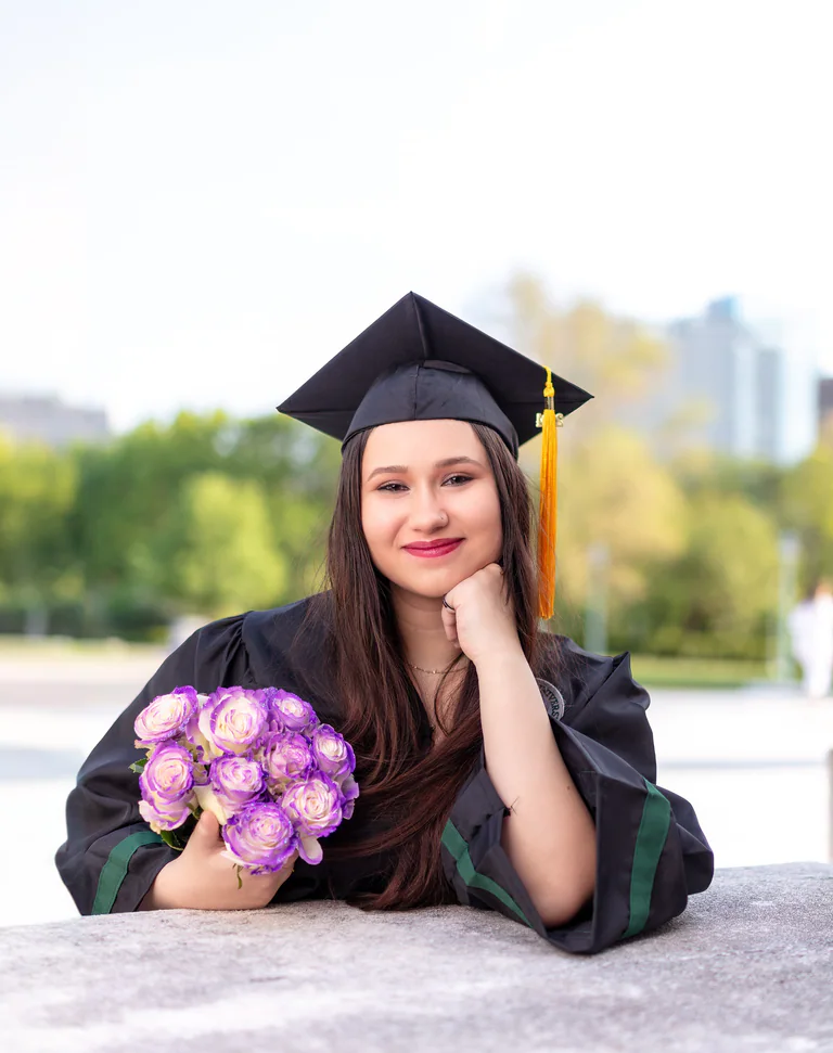 A graduate rests her cheek on one hand while holding a bouquet of purple flowers.