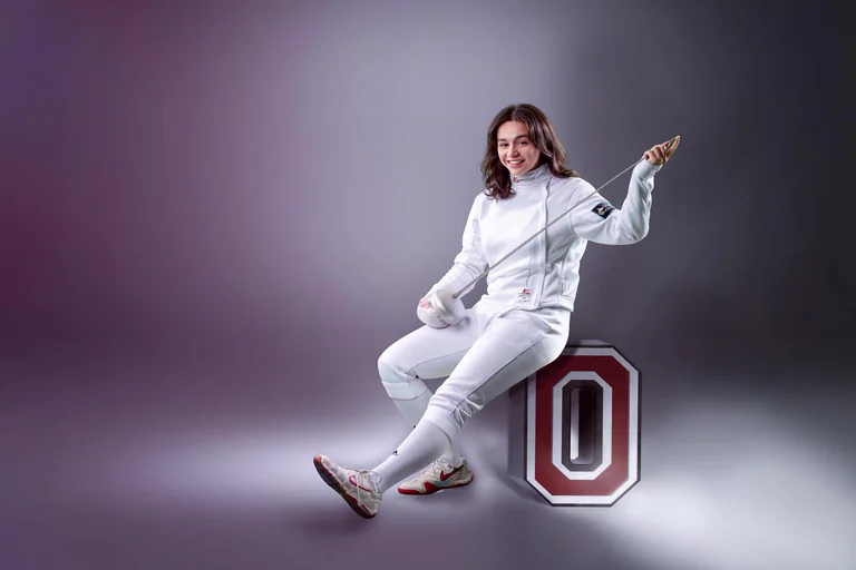 A fencer in white uniform poses beside a large block letter O against a purple background.