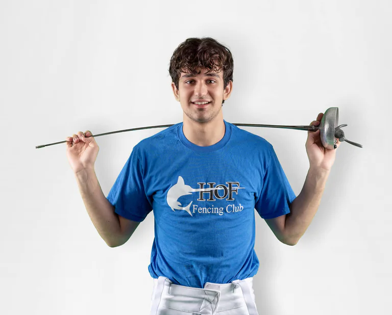 A smiling fencer in a blue shirt holds a foil across his shoulders and a fencing mask in one hand.