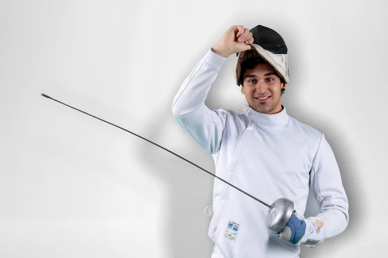 A fencer in a white jacket raises one hand while holding a foil against a pale background.