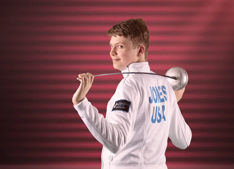 Rear portrait of a fencer holding a foil across one shoulder, with lettering on a white jacket.