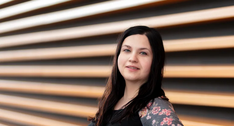 Portrait of a woman beside horizontal wooden slats, with soft light across her face.