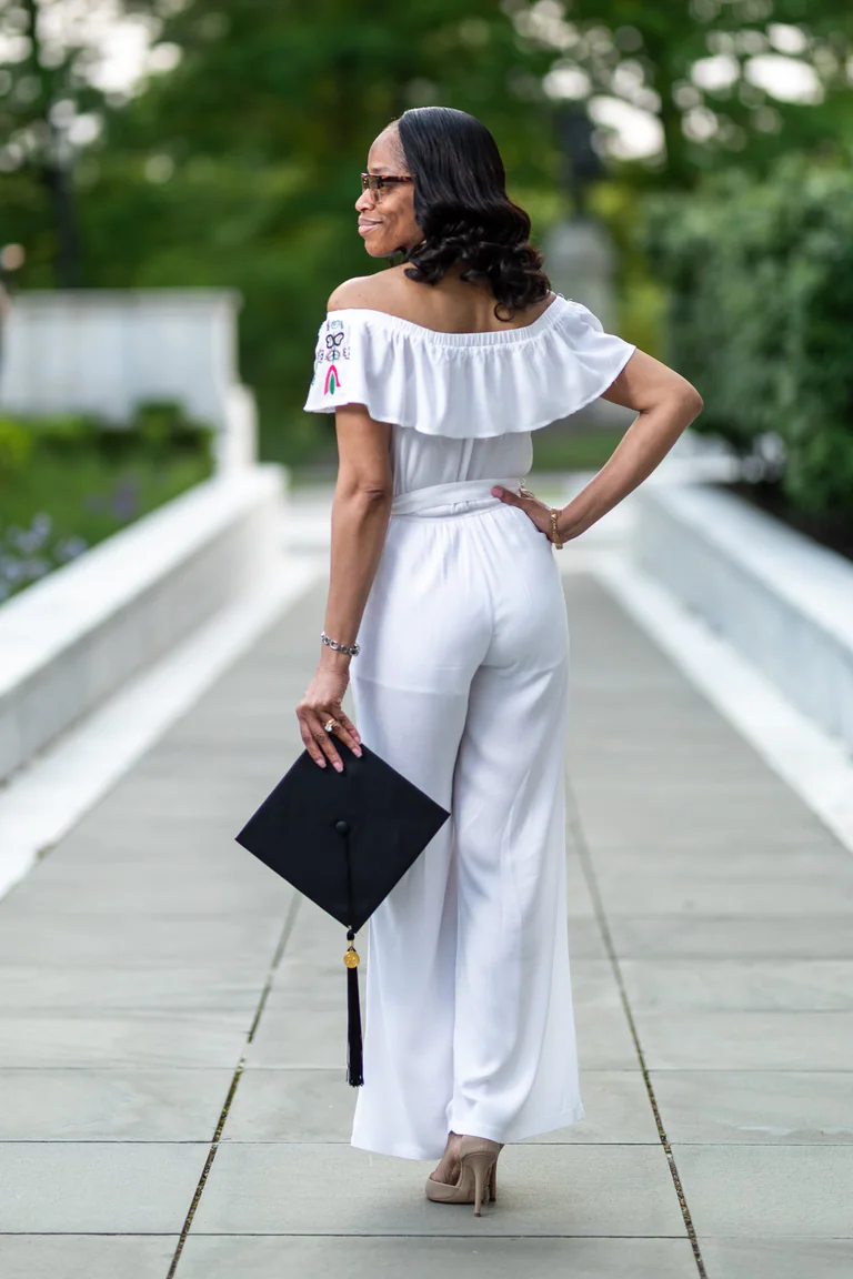 A woman in white walks along a wooden bridge, looking back while carrying a graduation cap.