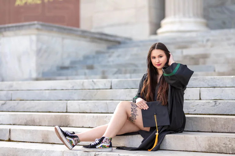 A woman in a graduation gown sits on broad stone steps with a mortarboard in her lap.