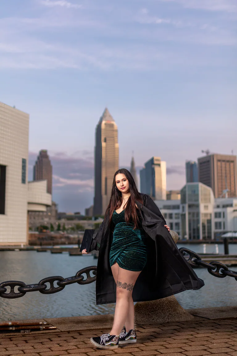 A woman poses beside a waterfront chain with tall city buildings rising across the water.