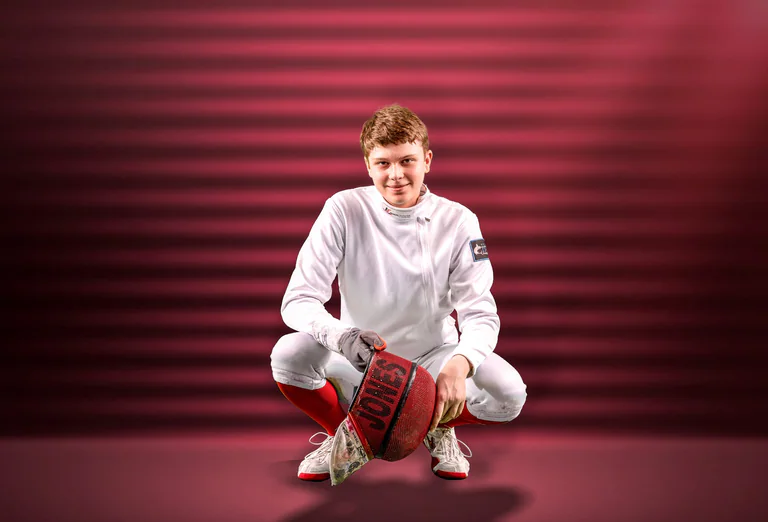 A fencer in white uniform crouches beside a red fencing mask against a striped red background.