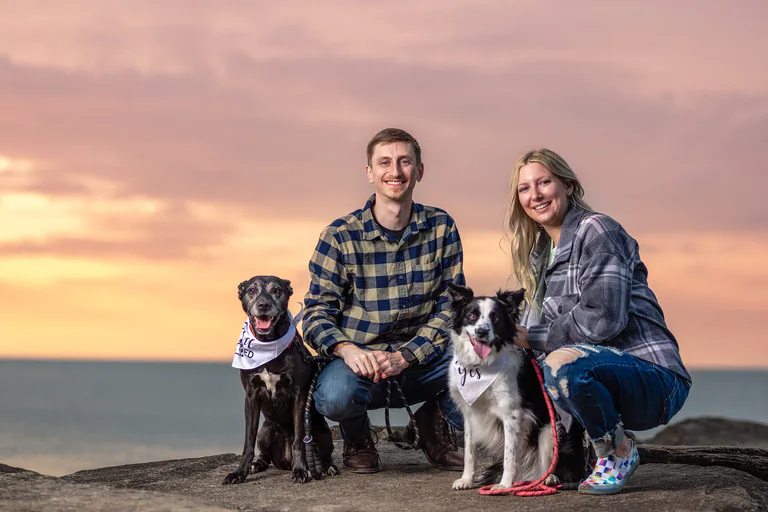 Two people pose with their dogs on a rocky overlook beneath a pink sunset sky.