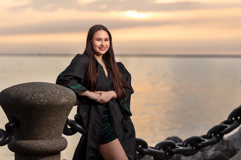 A woman in a dark outfit leans beside a mooring bollard and heavy chain at a waterfront sunset.