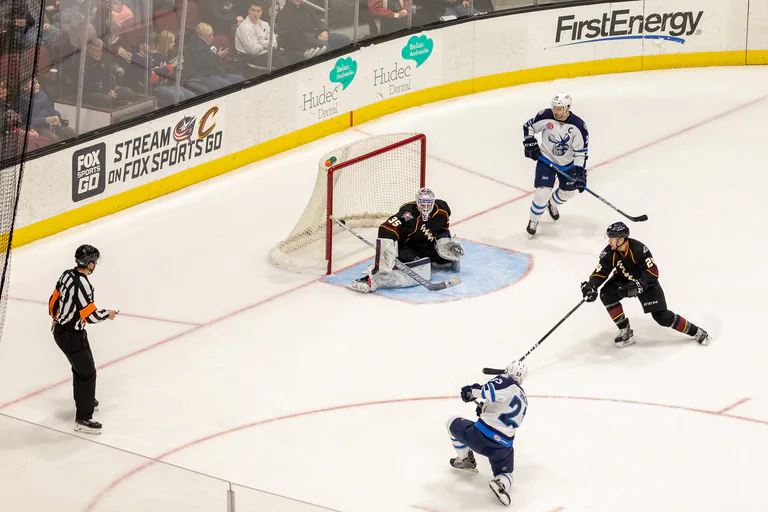 A black-uniformed goaltender reaches low while white-uniformed players approach the net.