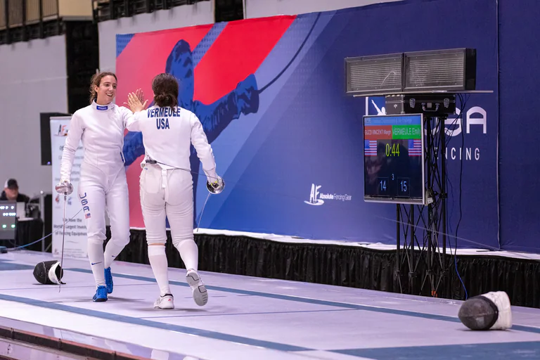 Two fencers stand together on the piste beside the scoring display.
