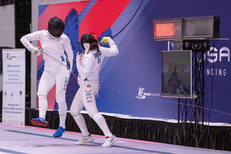 A fencer raises both arms while another stands beside the scoring display.