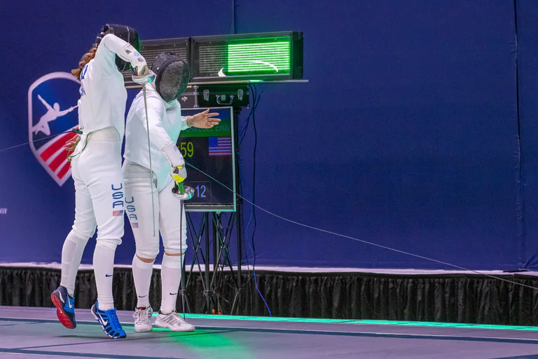 Two masked fencers meet at close range beside a green scoring light.