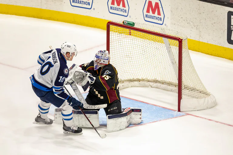 A player in white challenges a black-uniformed goaltender at the crease.