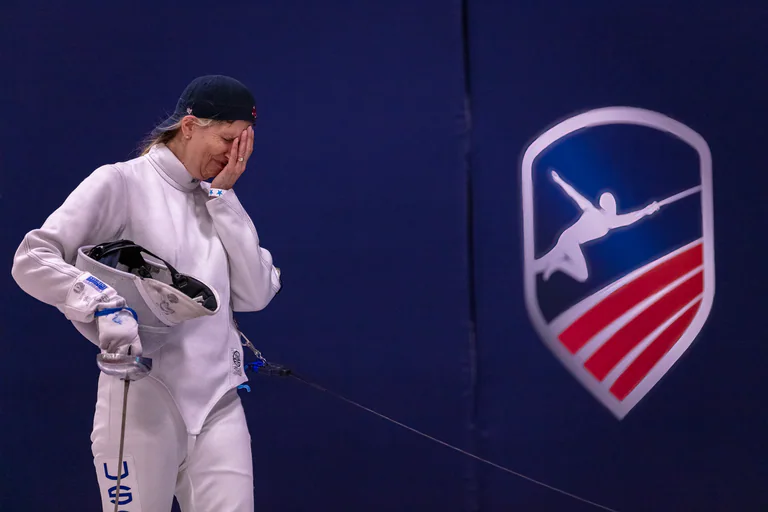 A fencer stands with mask removed and fencing equipment held at the hip.