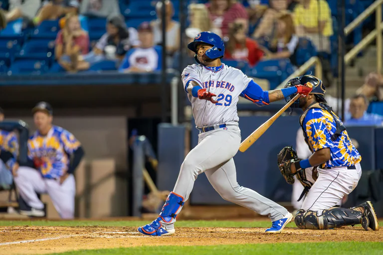A batter in white rotates through a swing as the catcher watches from behind.