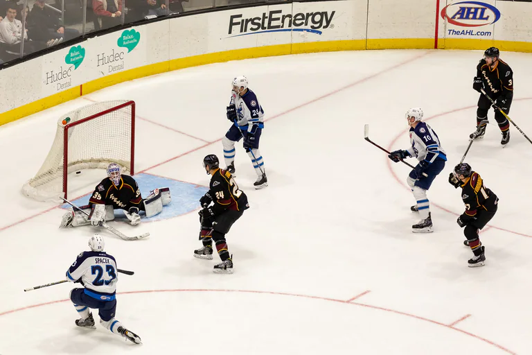 Hockey players gather around the goal as the goaltender stretches across the crease.
