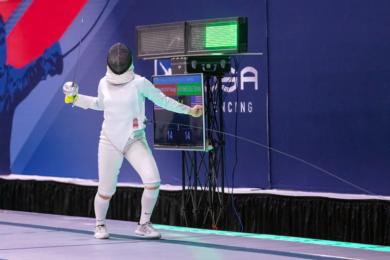 A masked fencer stands on the piste with both arms extended.
