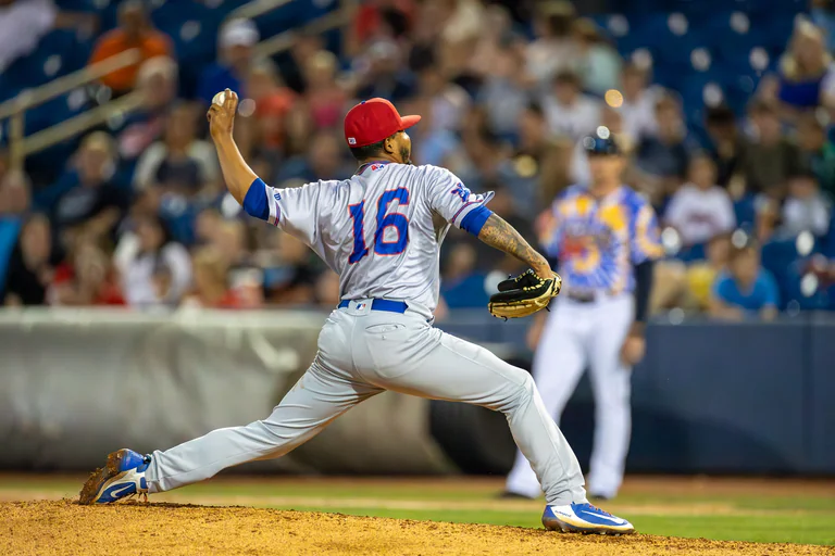 A pitcher in white drives forward from the mound with the throwing arm extended.