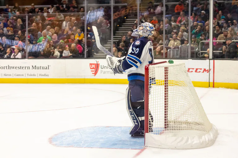 A blue-uniformed hockey goaltender stands watch beside the goal.